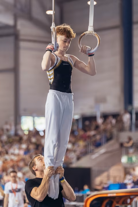 Young male gymnast holds still rings with focused expression while coach steadies his legs in packed gymnastics hall.