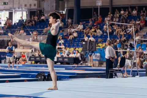 Male gymnast holds a precise arabesque balance on the blue floor mat, focused expression, during a gymnastics competition.