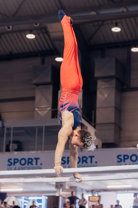 Male gymnast performs a perfect handstand on parallel bars, body fully extended upward, wearing red and navy leotard.