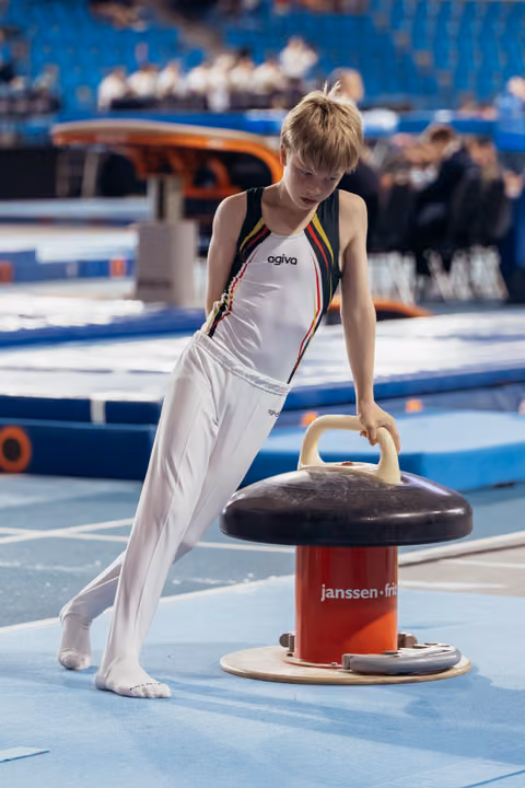 Young male gymnast in white leotard grips pommel horse handles, focused expression, blue gymnasium floor.