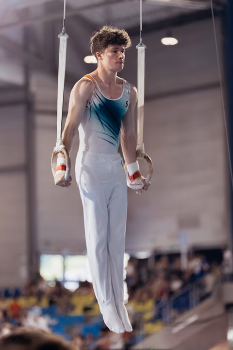 Young male gymnast hangs on still rings with intense focus, wearing teal leotard and white pants in a packed arena.
