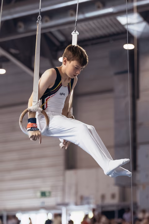 Focused young male gymnast holds a still rings position with intense concentration in a competition hall.