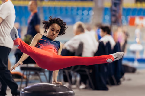 Young gymnast with curly hair performs a focused straddle hold on pommel horse, legs fully extended, intense concentration.