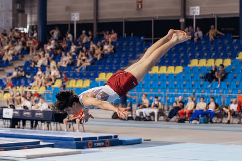 Young male gymnast executes a dramatic dive roll on floor, body fully extended, in a packed indoor arena.