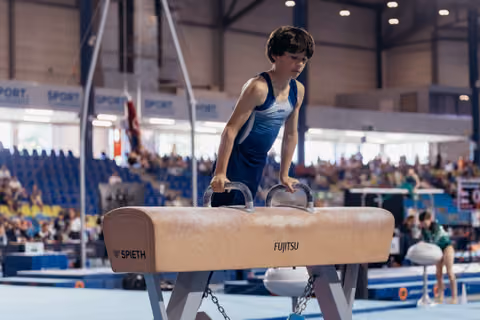 Young male gymnast grips pommel horse handles with intense focus during a gymnastics meet in a packed arena.