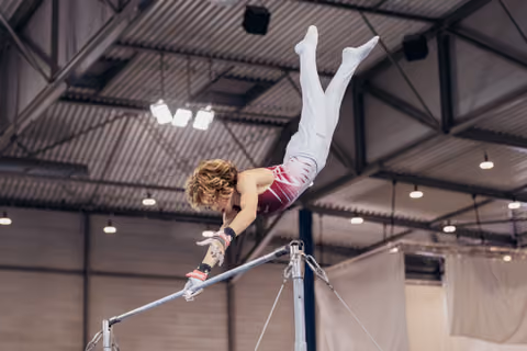 Young male gymnast releases the horizontal bar mid-air, body fully extended upside-down in an indoor gymnastics arena.