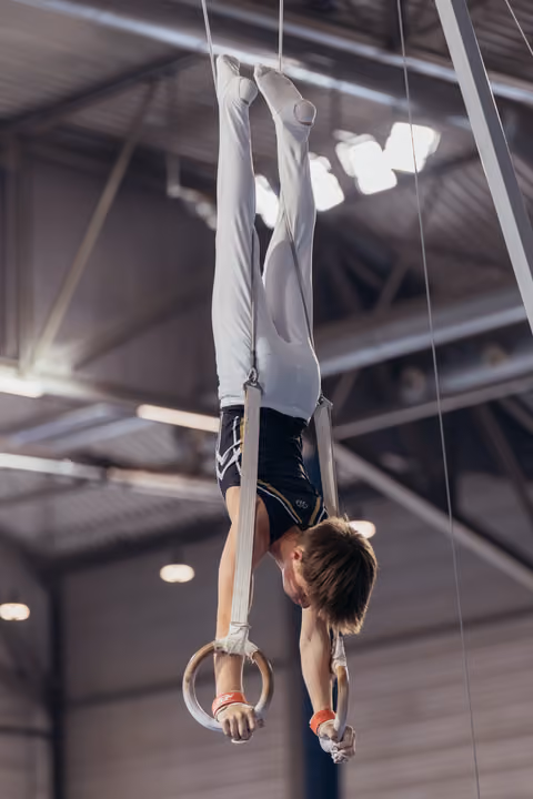 Young male gymnast performs an inverted hang on still rings, body fully extended, focused expression, in an indoor gymnasium.
