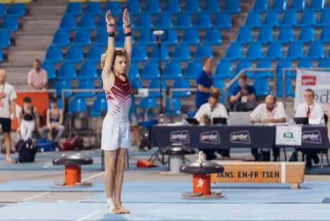 Young gymnast raises arms overhead in a focused salute pose on the floor exercise mat at a Gymfed competition.