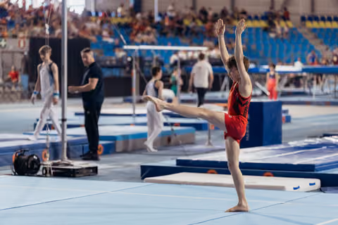 Young male gymnast in red leotard performs a high leg raise on the floor exercise mat, arms raised with focus.
