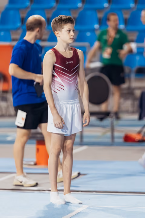 Young male gymnast in red and white Quatro leotard stands attentively on the floor, calm and focused, awaiting his turn.