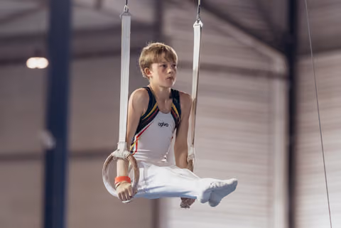 Young male gymnast grips still rings mid-routine, legs tucked, expression focused and determined in an indoor gym.