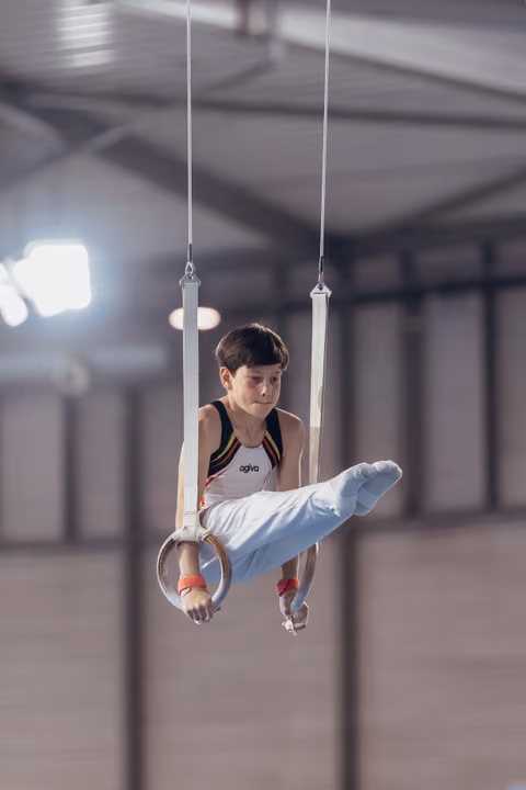Young male gymnast holds a tucked position on still rings, focused expression, competing indoors under bright lights.