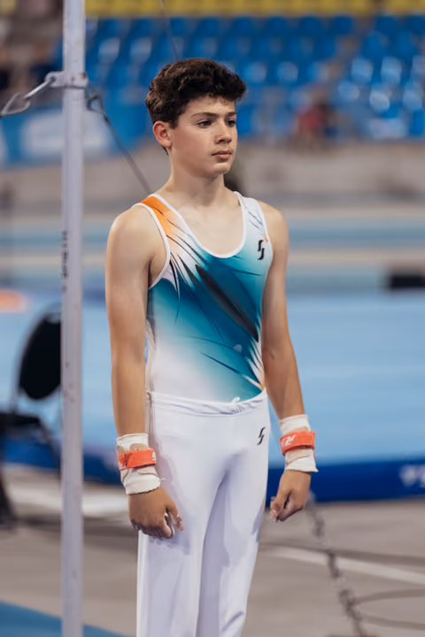 Young male gymnast stands focused and composed beside the high bar, wearing teal leotard with wrist guards, in a competition arena.