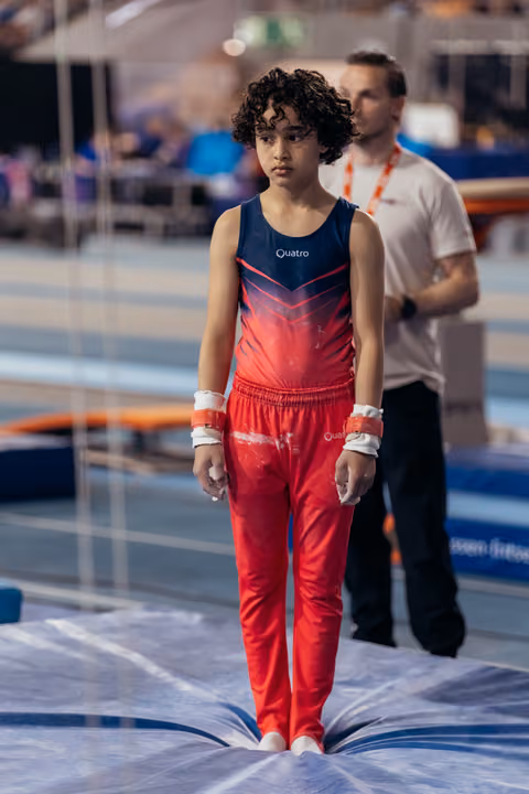 Young gymnast in red and navy Quatro leotard stands still on mat, expression serious and focused before performance.