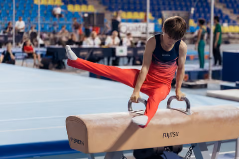 Young male gymnast performs focused pommel horse routine, legs extended, wearing red and navy at an indoor gymnastics meet.