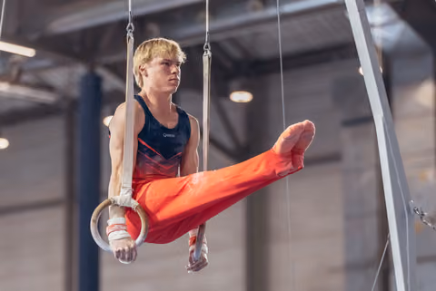Focused young male gymnast swings on still rings, leg raised, wearing red and navy leo in a gymnasium.