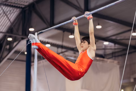 Young gymnast in red leotard hangs from horizontal bar with focused expression, legs extended in a straddle position.