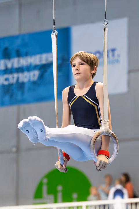 Young athlete performing on still rings with focused expression, wearing navy leotard with white pants and red wristbands