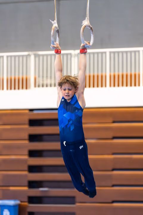 Young athlete hanging from rings with focused expression during training, wooden bleachers visible in background