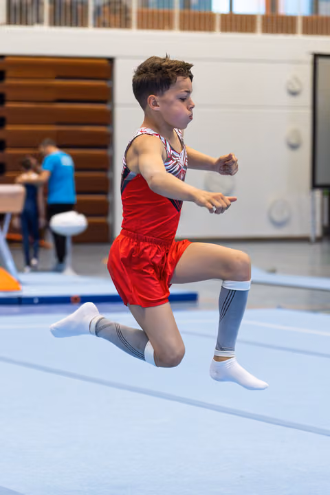 Young athlete jumping mid-air during floor exercise routine in gymnasium, wearing red shorts and striped leotard