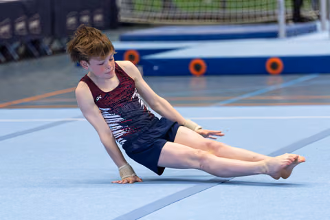 Young athlete performs seated floor routine with extended legs on blue mat at indoor gymnastics facility
