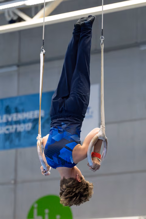 Athlete performs an inverted hang on still rings, displaying strength and control with legs extended upward in an indoor gym