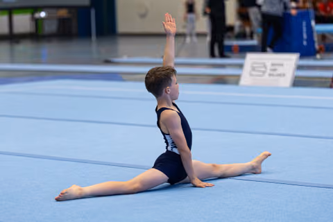 Young gymnast performs a split with one arm raised during floor exercise at indoor sports competition
