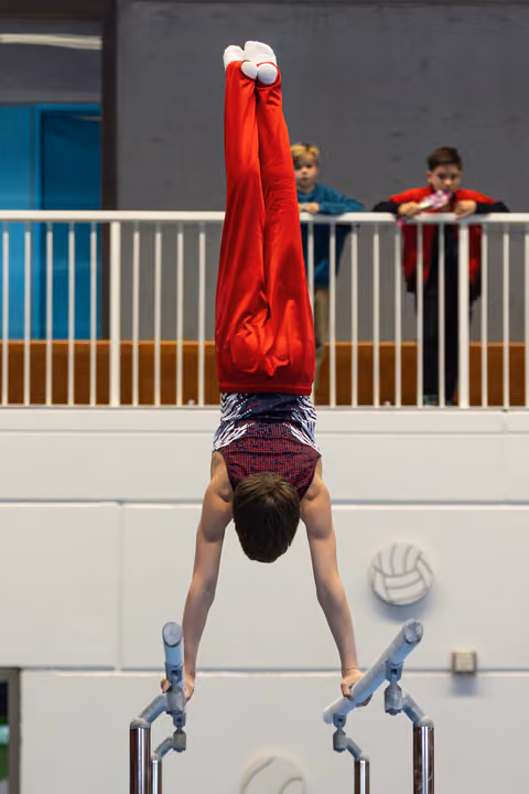Young athlete performing a handstand on parallel bars with pointed toes, wearing red shirt, while spectators watch from balcony