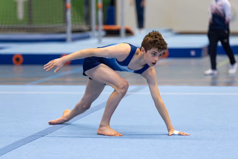 Young athlete in navy leotard strikes a dramatic starting pose on floor mat, displaying intense focus and athletic form