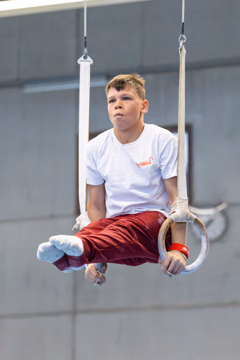Athlete performing still rings hold, focused expression, white shirt and burgundy pants, indoor training facility