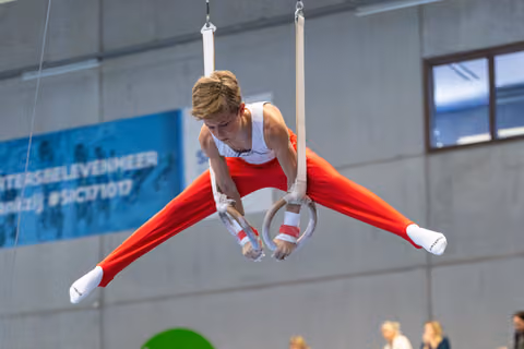 Young athlete performs straddle position on rings, legs extended horizontally, wearing red and white uniform at indoor event