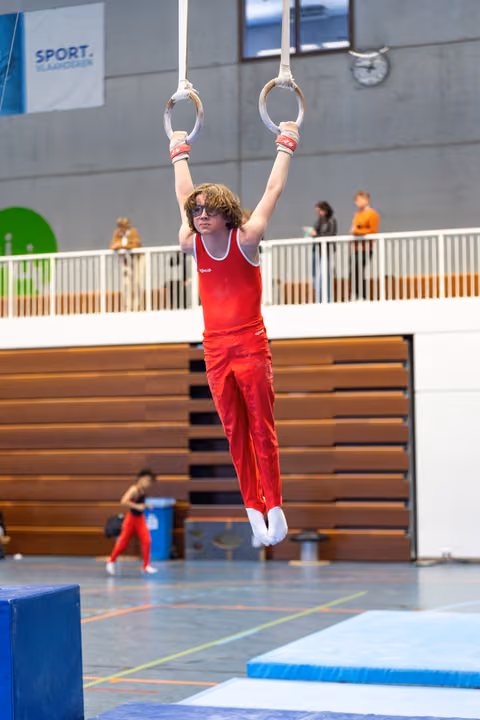 Young athlete in red uniform hanging from rings mid-air, displaying strength and focus during routine at indoor sports facility