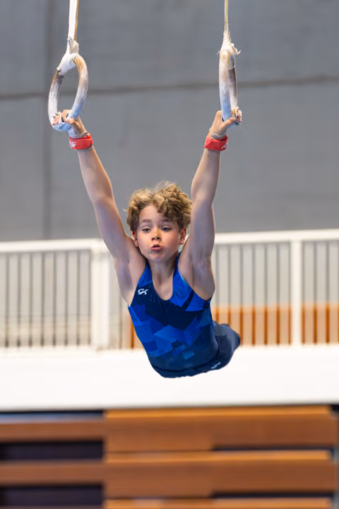 Young athlete performs a hang on still rings with extended arms, wearing red grips and blue leotard, focused expression