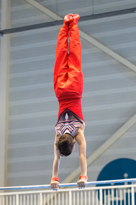 Athlete performing a handstand on parallel bars with straight vertical alignment in red tracksuit at indoor training facility
