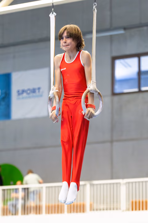 Young athlete suspended on still rings in red uniform, showing focused concentration during routine at indoor training facility