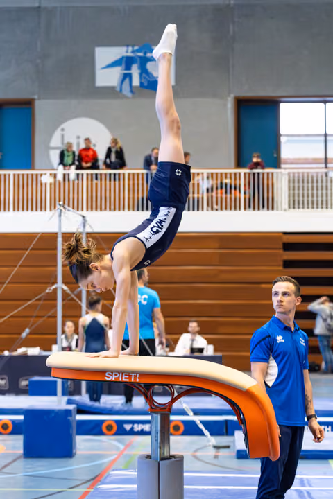 Athlete performs a handstand on vault apparatus while coach spots nearby during indoor sports event