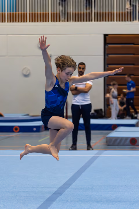 Young athlete performs a graceful leap on floor exercise, arms extended, with coach observing in background