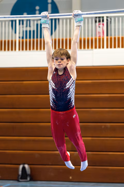Young gymnast hangs from horizontal bar with arms extended overhead in red and black leotard during training session