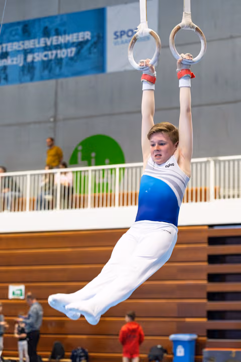 Young gymnast hanging from still rings in white and blue uniform, demonstrating strength hold in indoor sports hall