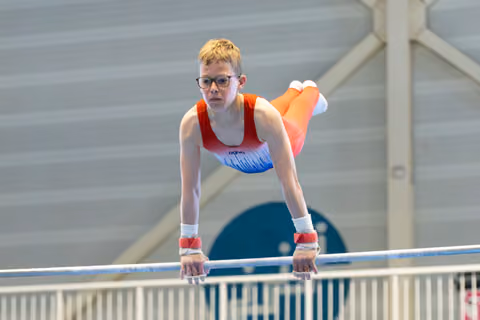 Young gymnast with blonde hair and glasses performs on horizontal bar with intense concentration, wearing red and blue leotard