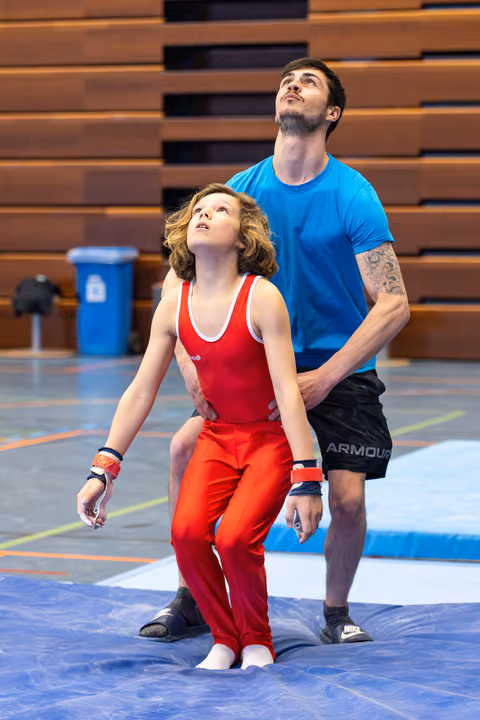 Coach guiding young athlete in red singlet, both looking upward during training session in indoor sports hall