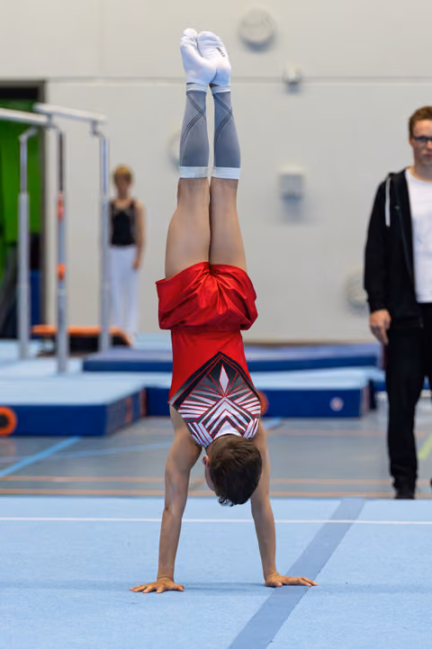 Young athlete performs a handstand on the floor mat, wearing a red leotard and blue shorts, with coaches observing