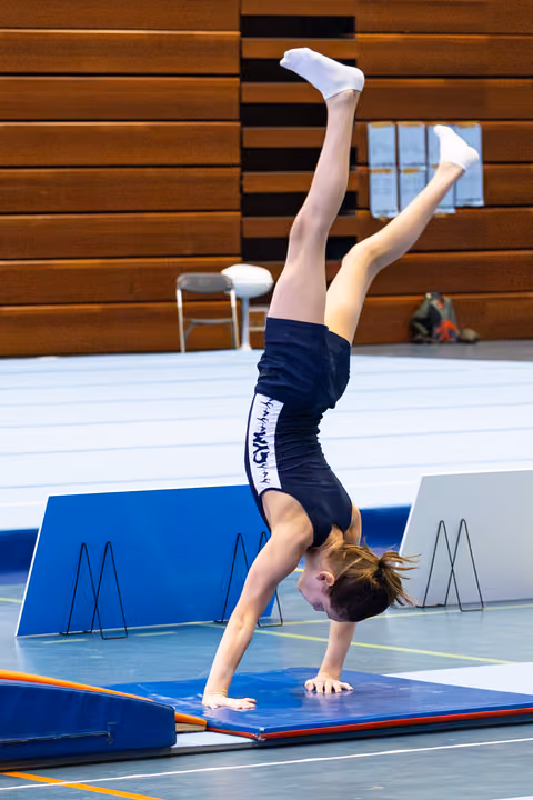 Young athlete performing a handstand on blue training mat in gymnasium, legs extended upward, wearing navy uniform