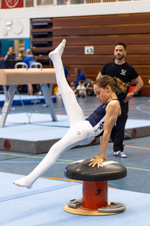 Female gymnast executing a horizontal split on pommel horse with legs extended vertically, coach observing in background