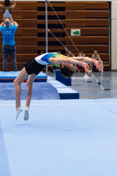 Young athlete executes a dramatic backbend mid-flight during floor exercise, arms extended backwards in indoor training facility