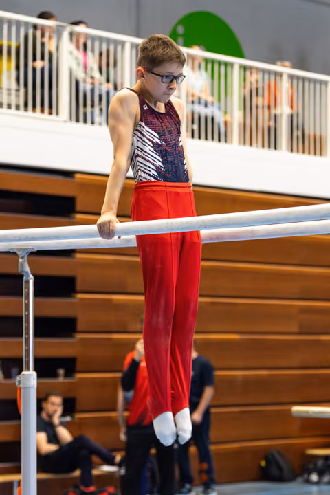 Young gymnast in red pants and patterned leotard performing on parallel bars with focused expression, spectators watching