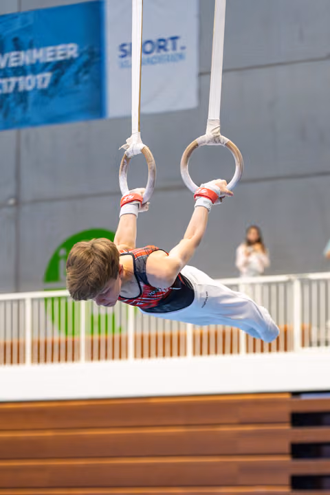 Young athlete performing an inverted hang on still rings during indoor competition, demonstrating upper body strength