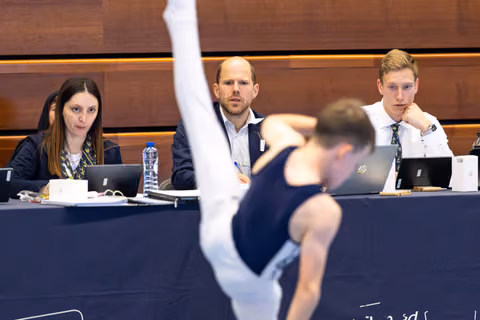 Three judges at table with laptops observe athlete mid-routine with focused expressions during evaluation