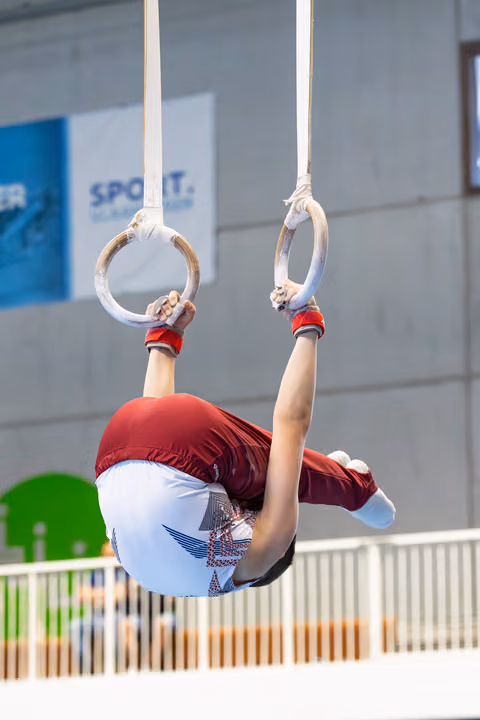 Athlete performing an inverted hang on still rings with legs tucked, wearing red and white uniform at indoor sports facility
