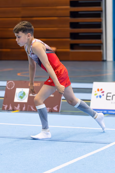 Young athlete in red shorts running across blue floor mat during routine, focused expression, wooden bleachers in background